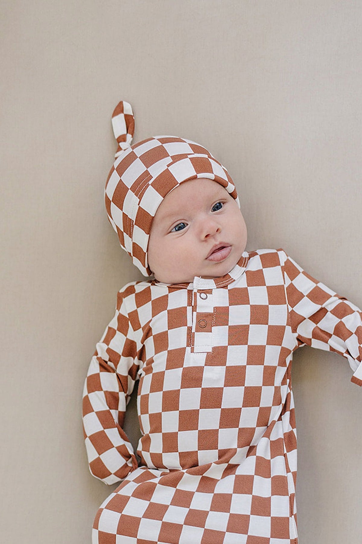 A newborn wears a rust checkered knot hat and matching outfit made of soft bamboo fabric. The baby is lying on a neutral-toned background, showcasing the stylish, gender-neutral design.
