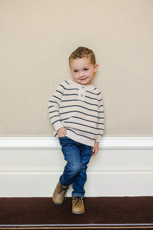A young child wearing a cream and navy striped sweater, jeans, and brown shoes stands against a beige wall.