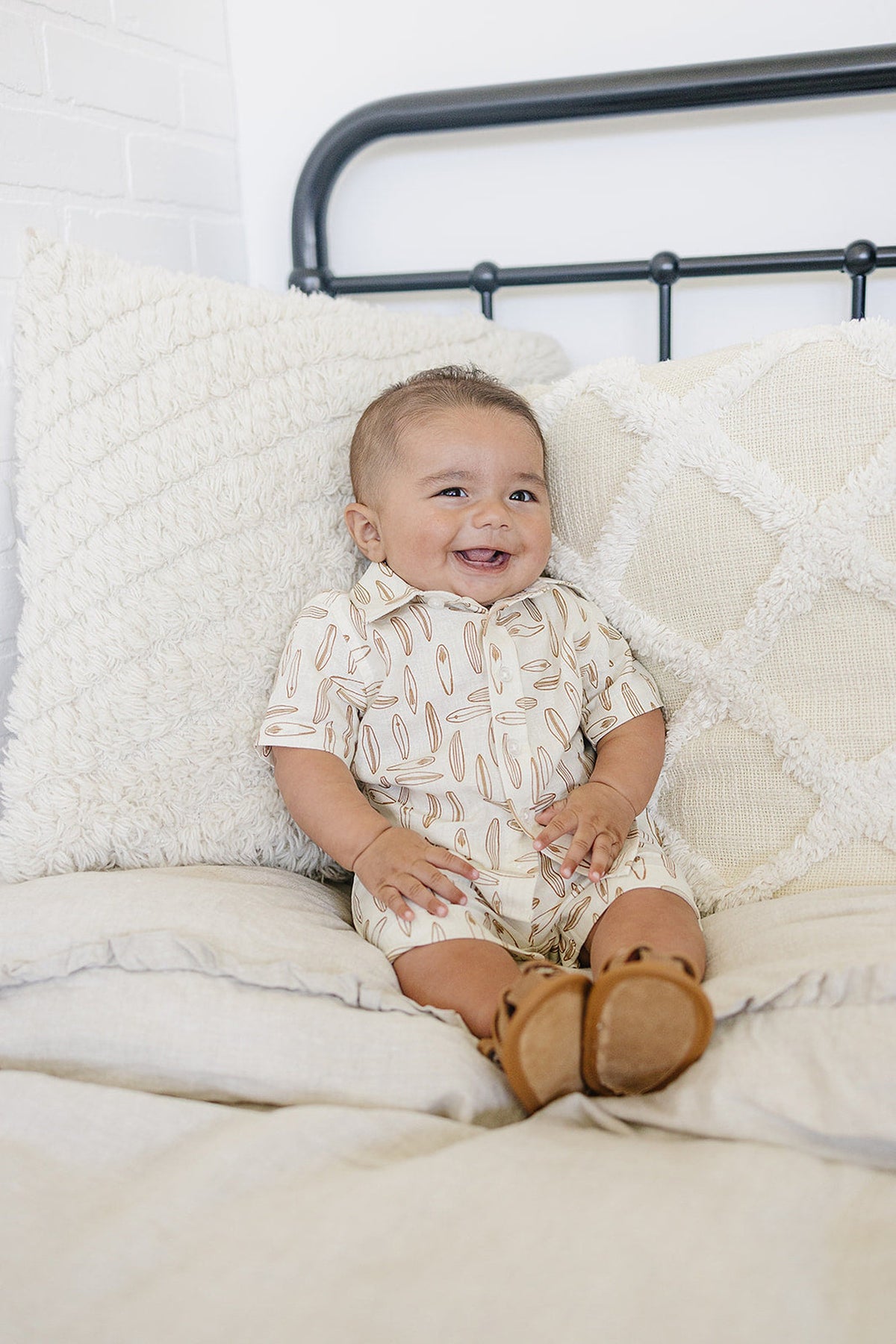 A smiling baby wearing a surfboard-patterned onesie sits on a bed with textured pillows.