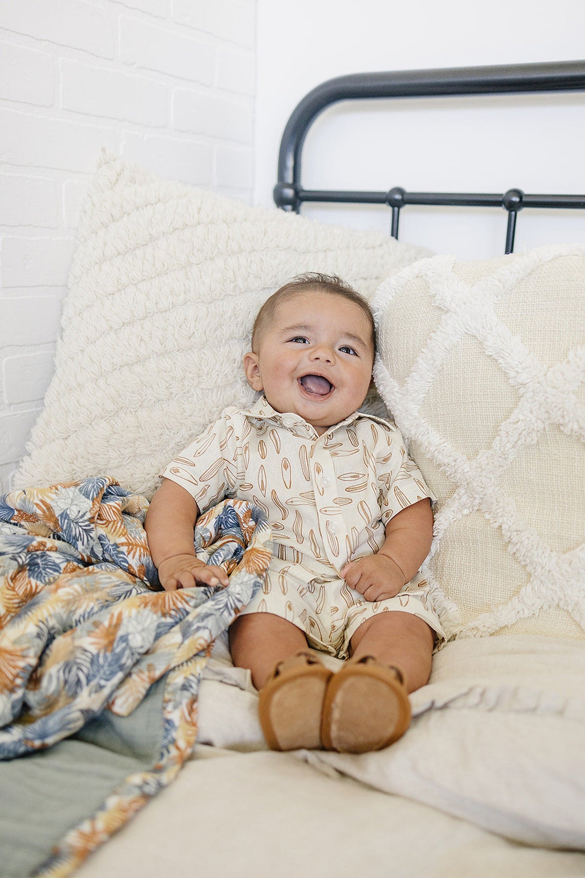 A smiling baby wearing a surfboard-patterned button-up shirt sits on a bed with textured pillows and a colorful blanket.