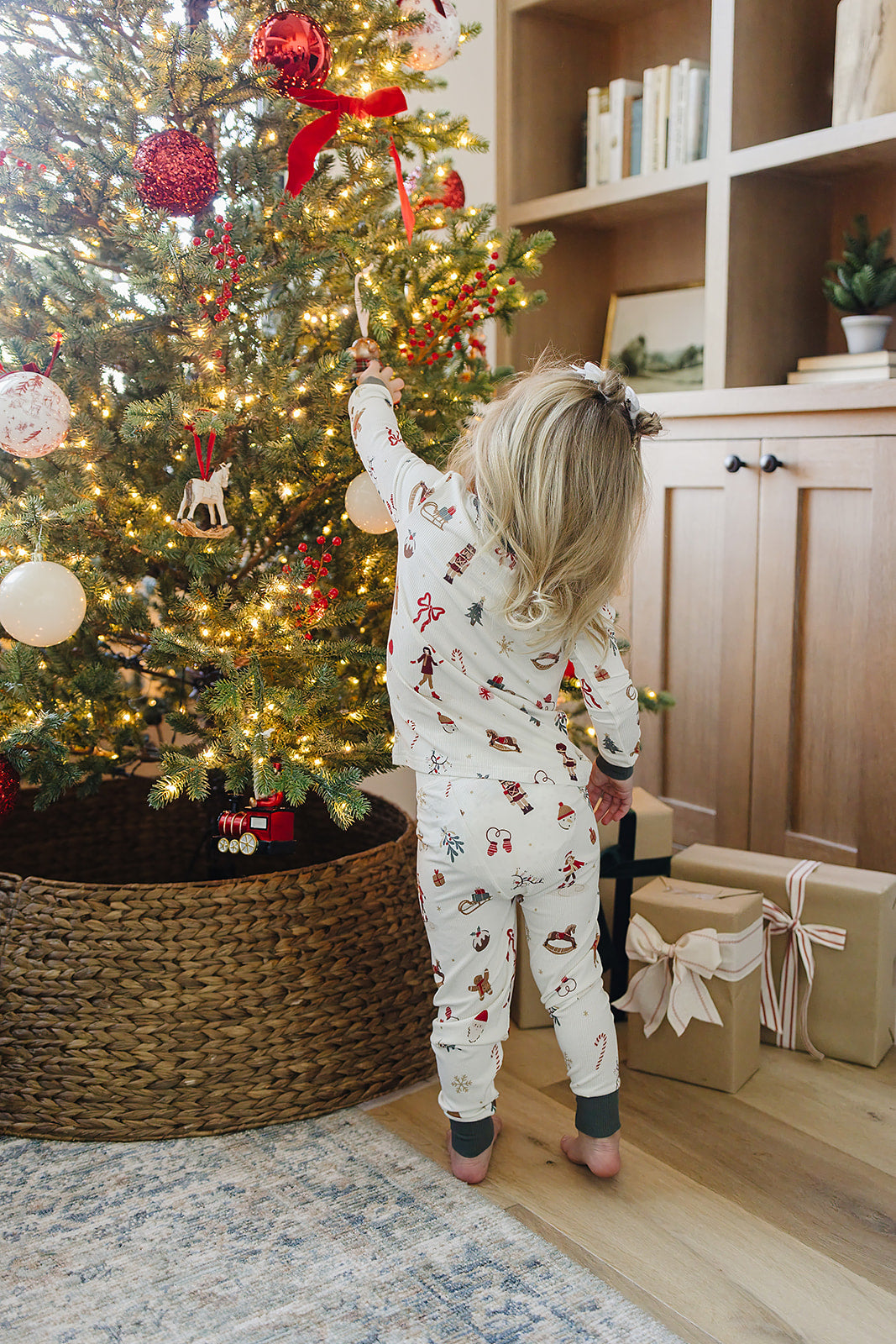 A child in festive pajamas reaches for an ornament on a decorated Christmas tree, surrounded by wrapped gifts. The cozy set features a cheerful holiday print and soft fabric.