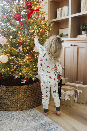 A child in festive pajamas reaches for an ornament on a decorated Christmas tree, surrounded by wrapped gifts. The cozy set features a cheerful holiday print and soft fabric.