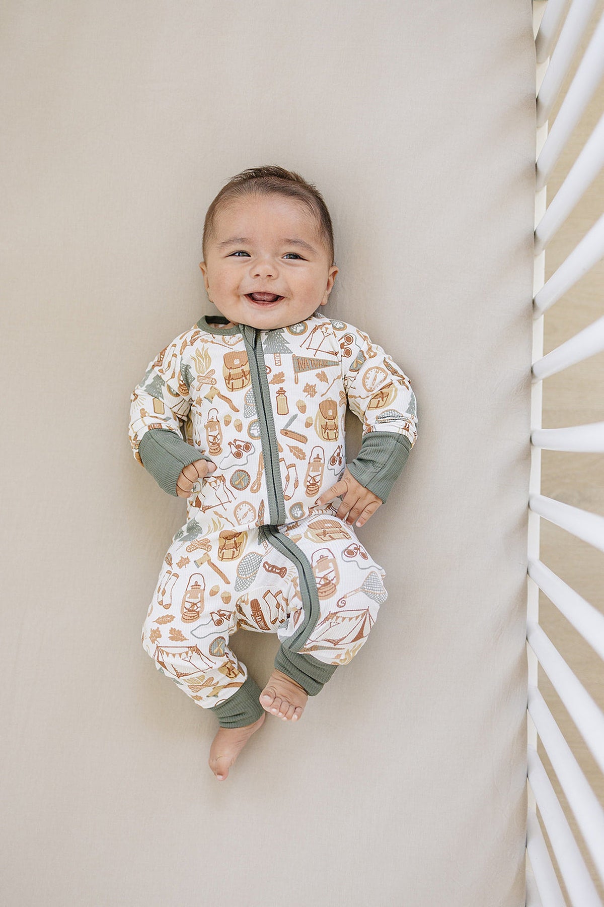 A smiling baby wears a camping-themed, bamboo zipper pajama with green cuffs, lying in a crib.