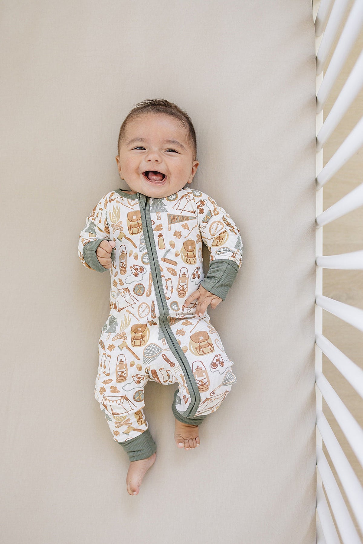 A smiling baby wears camping-themed, bamboo pajamas with green cuffs, lying in a crib on a beige sheet.