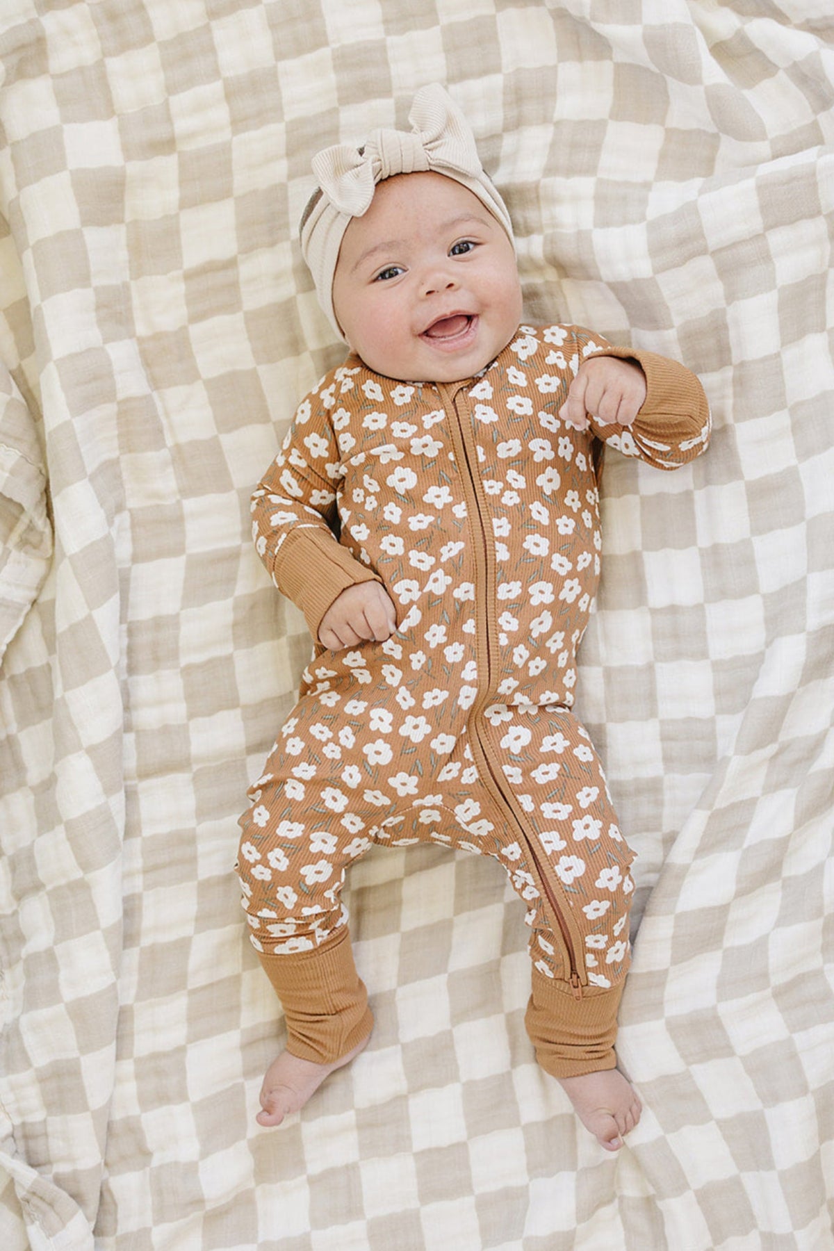 Smiling baby in floral mustard ribbed pajamas on a checkered blanket, wearing a cream bow headband.