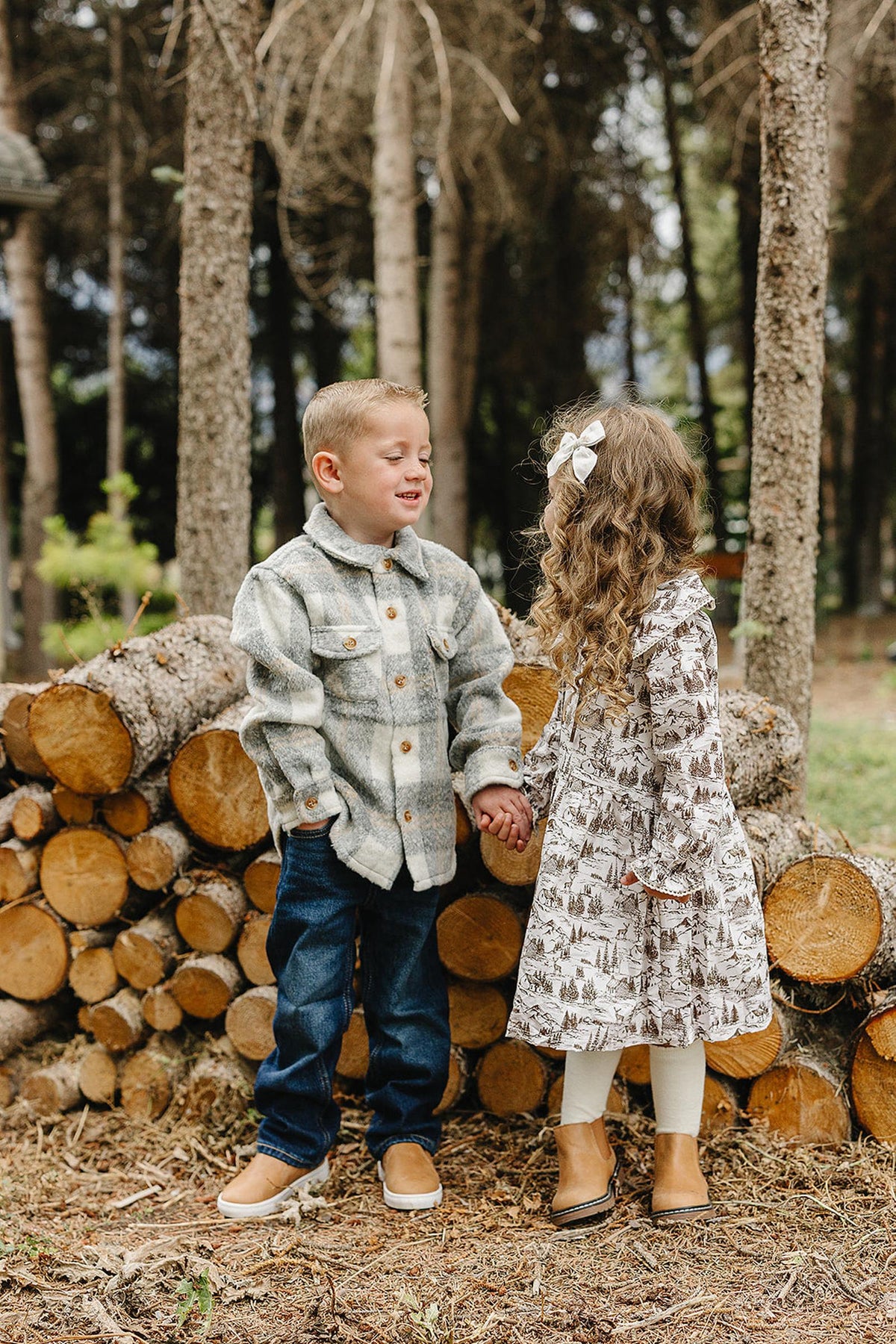 A young boy in a flannel shirt and jeans holds hands with a girl in a brown toile long-sleeve dress, standing among stacked logs in a forest setting.