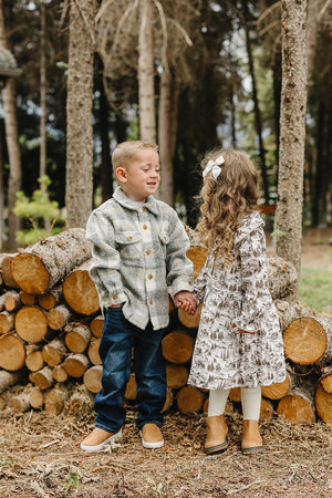 A young boy in a flannel shirt and jeans holds hands with a girl in a brown toile long-sleeve dress, standing among stacked logs in a forest setting.