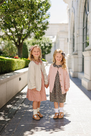 Two young girls wearing knit cardigans and floral dresses hold hands outdoors, surrounded by greenery and architecture.