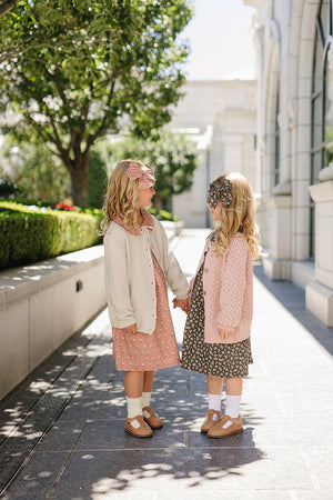 Two young girls holding hands, wearing floral dresses and cardigans, with matching headbands, standing outdoors near greenery.