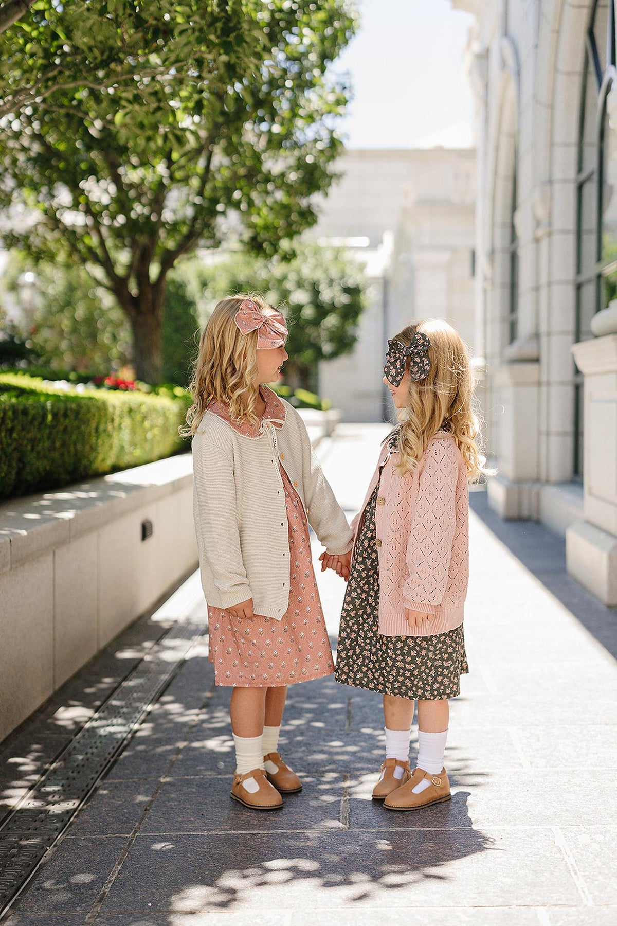 Two young girls wearing floral dresses and cardigans hold hands, each adorned with large fabric hair bows.
