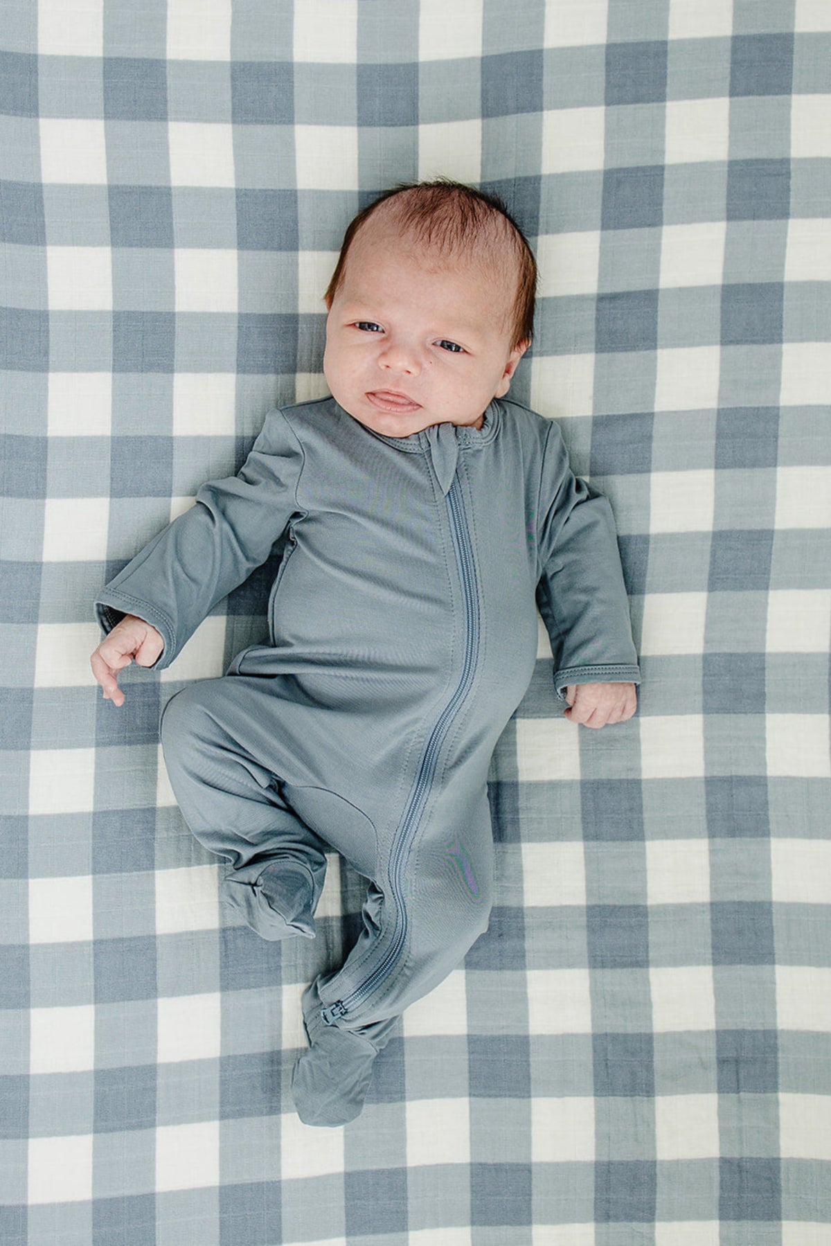 A baby in a blue onesie lies on a dusty blue gingham crib sheet made of soft muslin cotton.