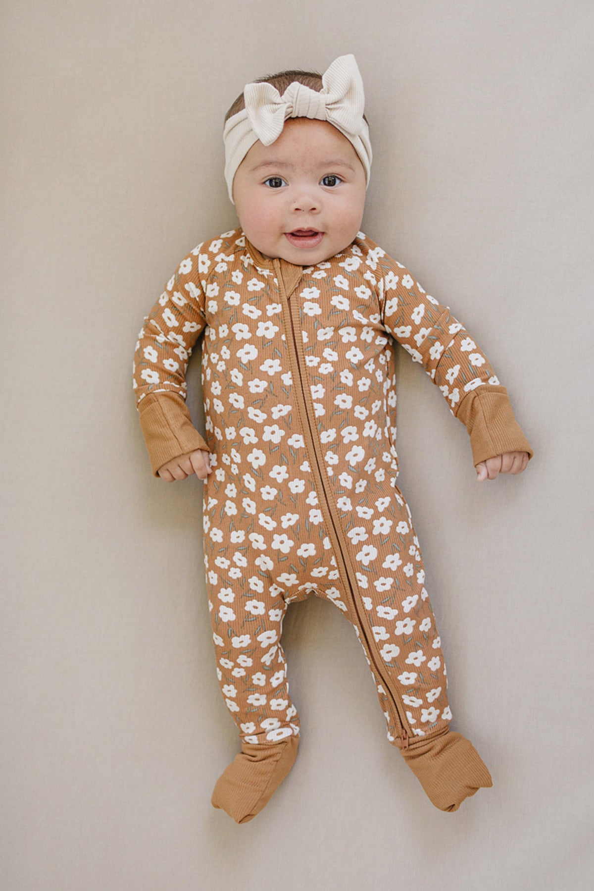 A baby wearing a mustard floral ribbed pajama and a beige bow headband, smiling on a light background.