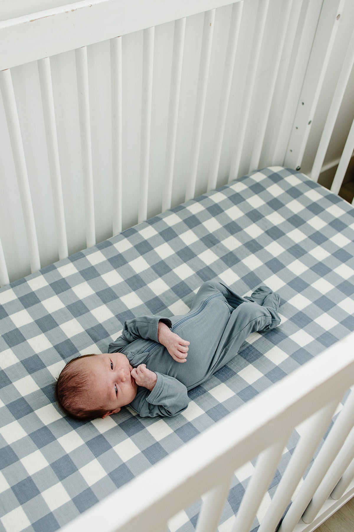 A baby in a blue onesie lies on a dusty blue gingham muslin crib sheet in a white crib.