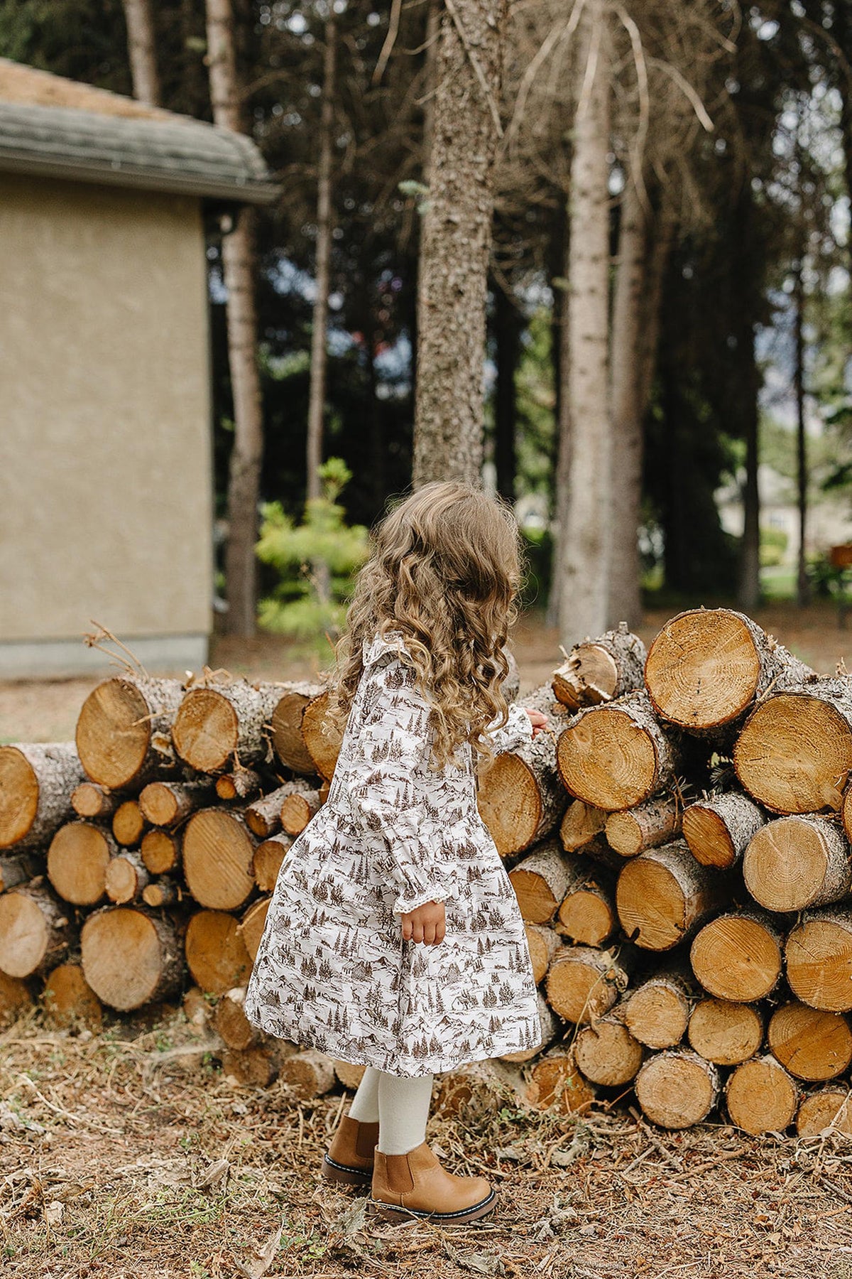 A young girl with curly hair wears a brown toile long-sleeve dress while exploring a stack of wooden logs in a natural outdoor setting.