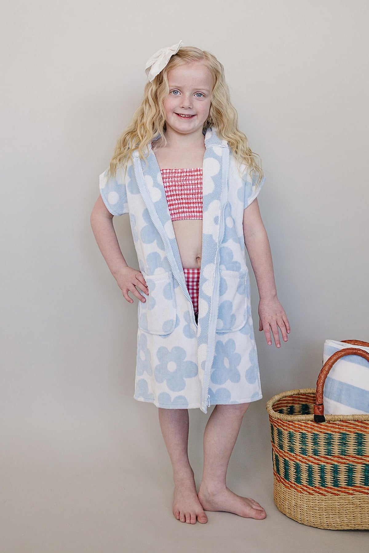A young girl poses in a light blue and white floral zip-up poncho made from soft organic cotton. She stands beside a woven basket, showcasing a playful, beach-ready look.