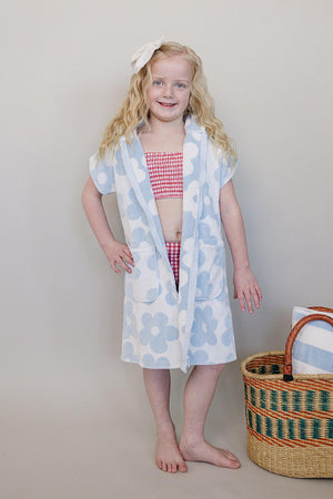 A young girl poses in a light blue and white floral zip-up poncho made from soft organic cotton. She stands beside a woven basket, showcasing a playful, beach-ready look.