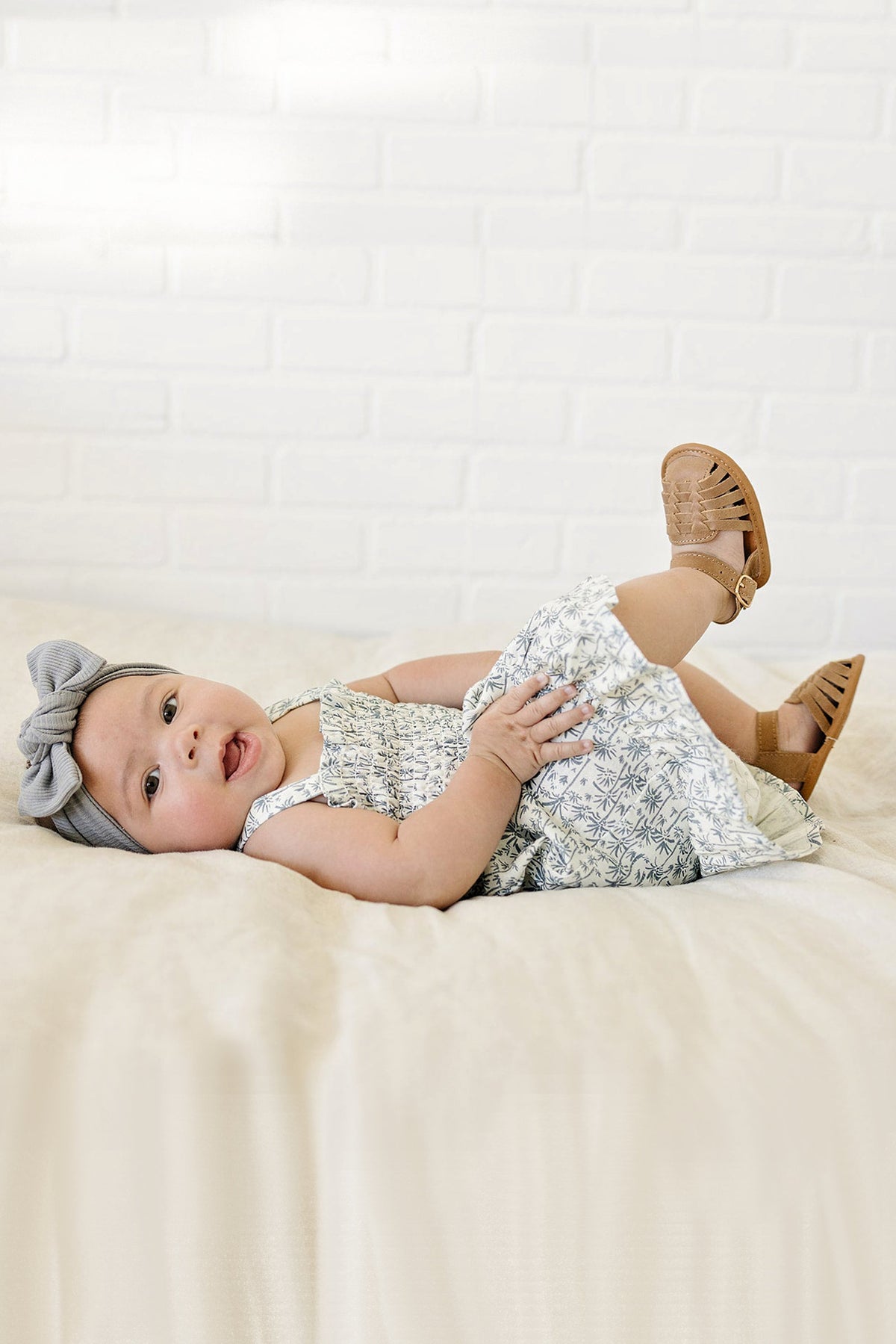 Baby wearing a palm tree print dress and a grey headband, lying on a soft beige surface.