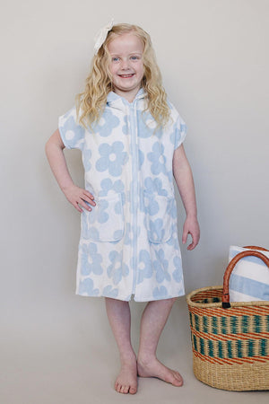 A young girl stands smiling, wearing a light blue and white floral zip-up beach poncho made of organic cotton. A woven basket sits beside her, adding a beachy vibe.
