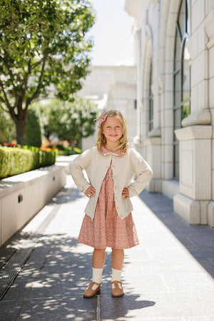 A young girl in a pink floral dress and cardigan poses outdoors, surrounded by greenery and stone architecture.