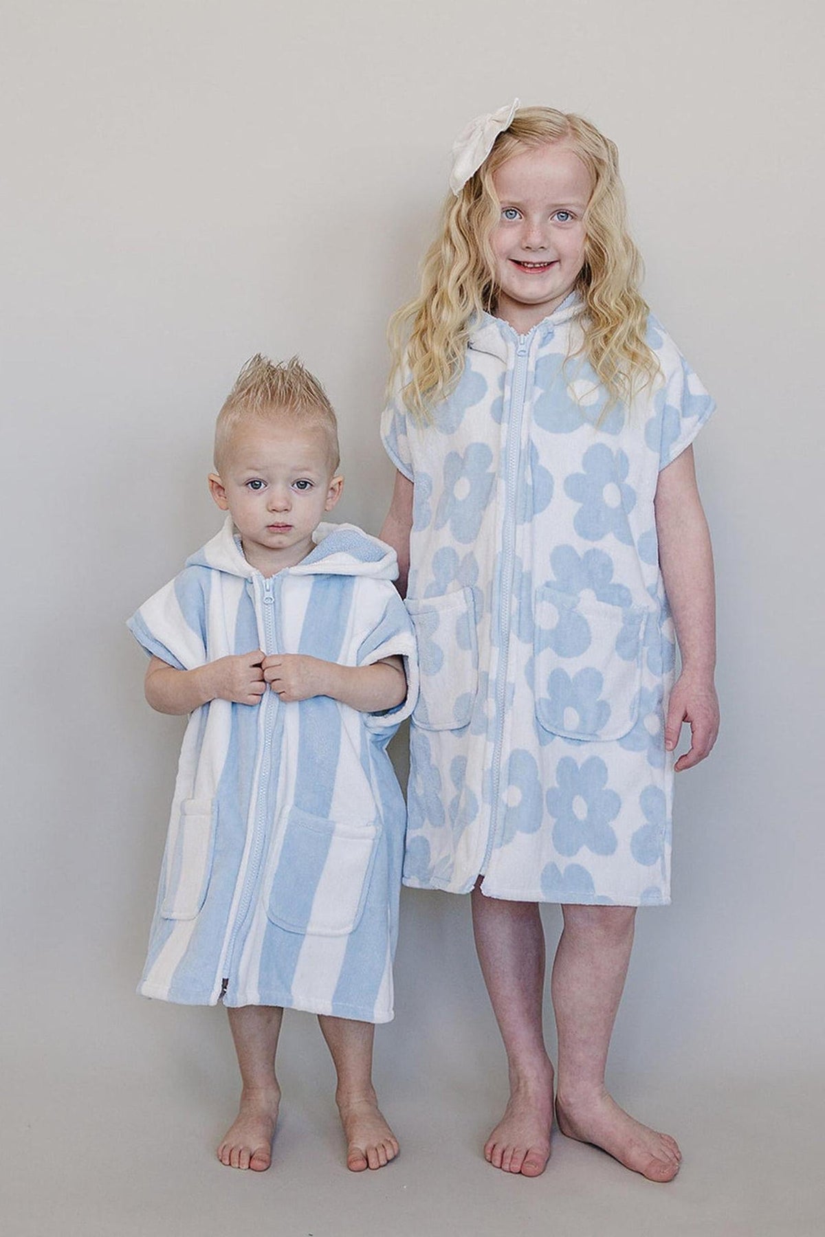 Two children stand side by side wearing light blue and white zip-up beach ponchos made from soft, breathable organic cotton. The ponchos feature floral patterns and convenient pockets.