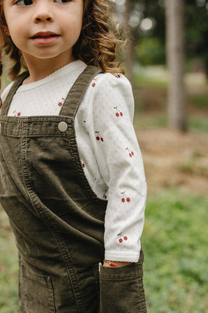 A young child wears a white long-sleeve shirt with cherry patterns, layered under a brown corduroy overall.