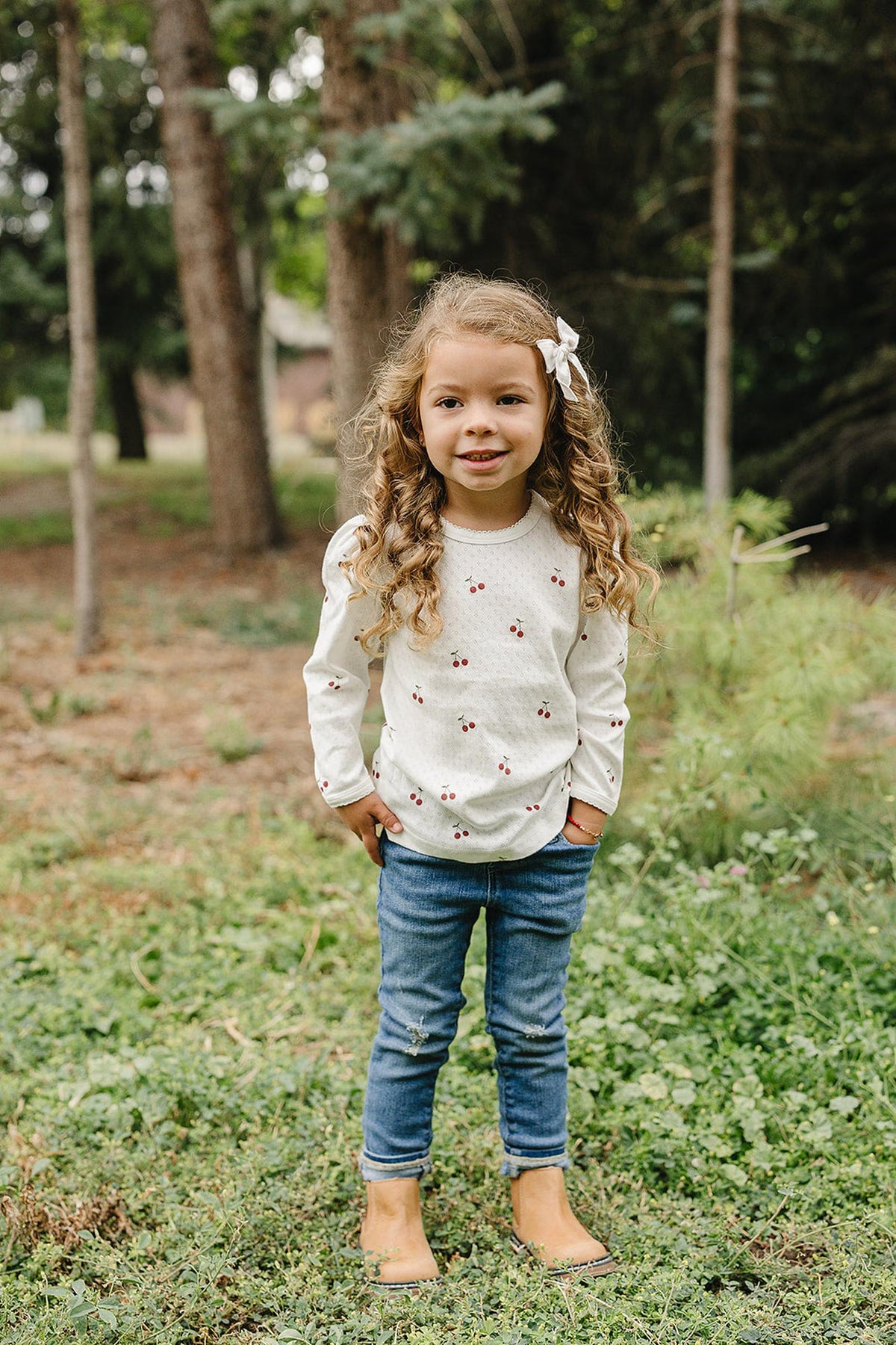 A young girl with curly hair stands in a grassy area, wearing a cream long sleeve shirt with cherry designs.