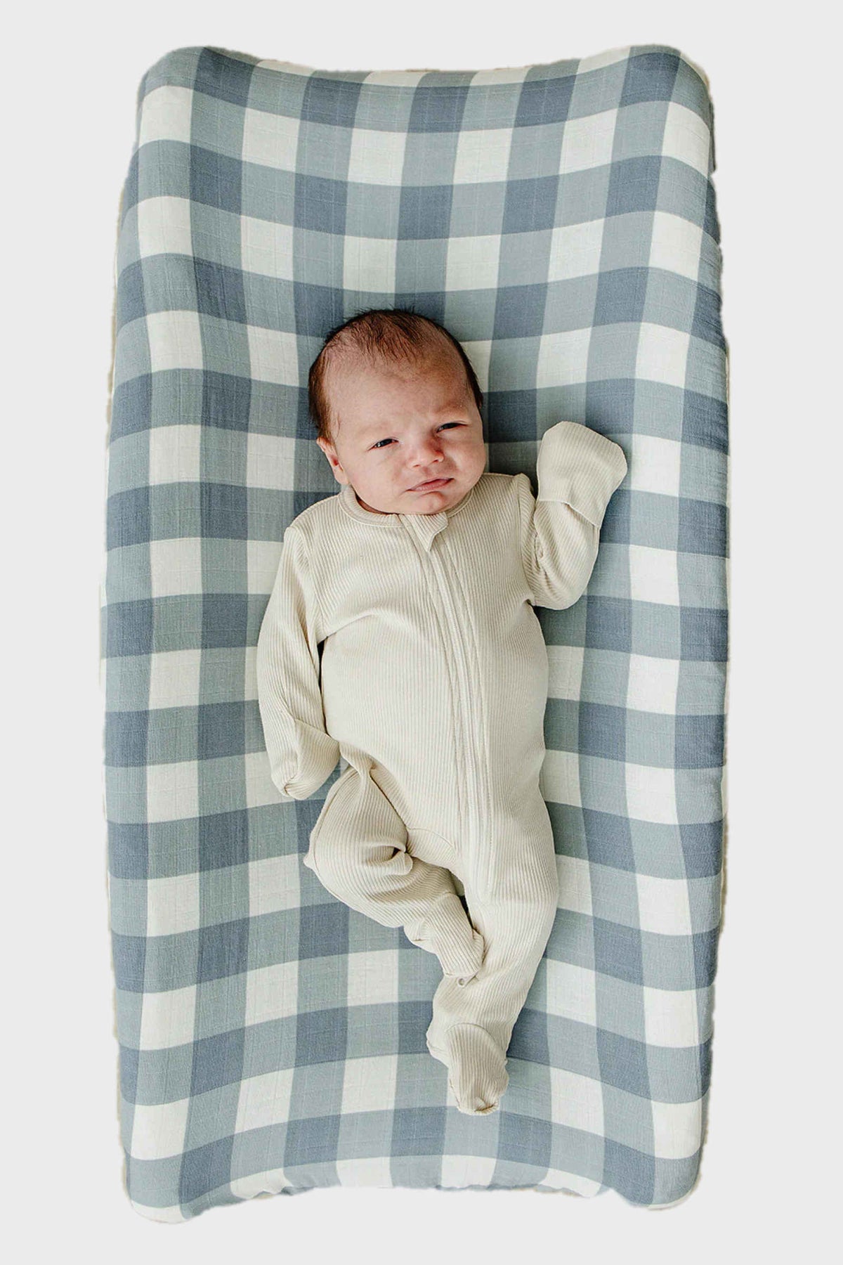 Baby in cream onesie lying on a dusty blue gingham muslin changing pad cover with a checkered pattern.