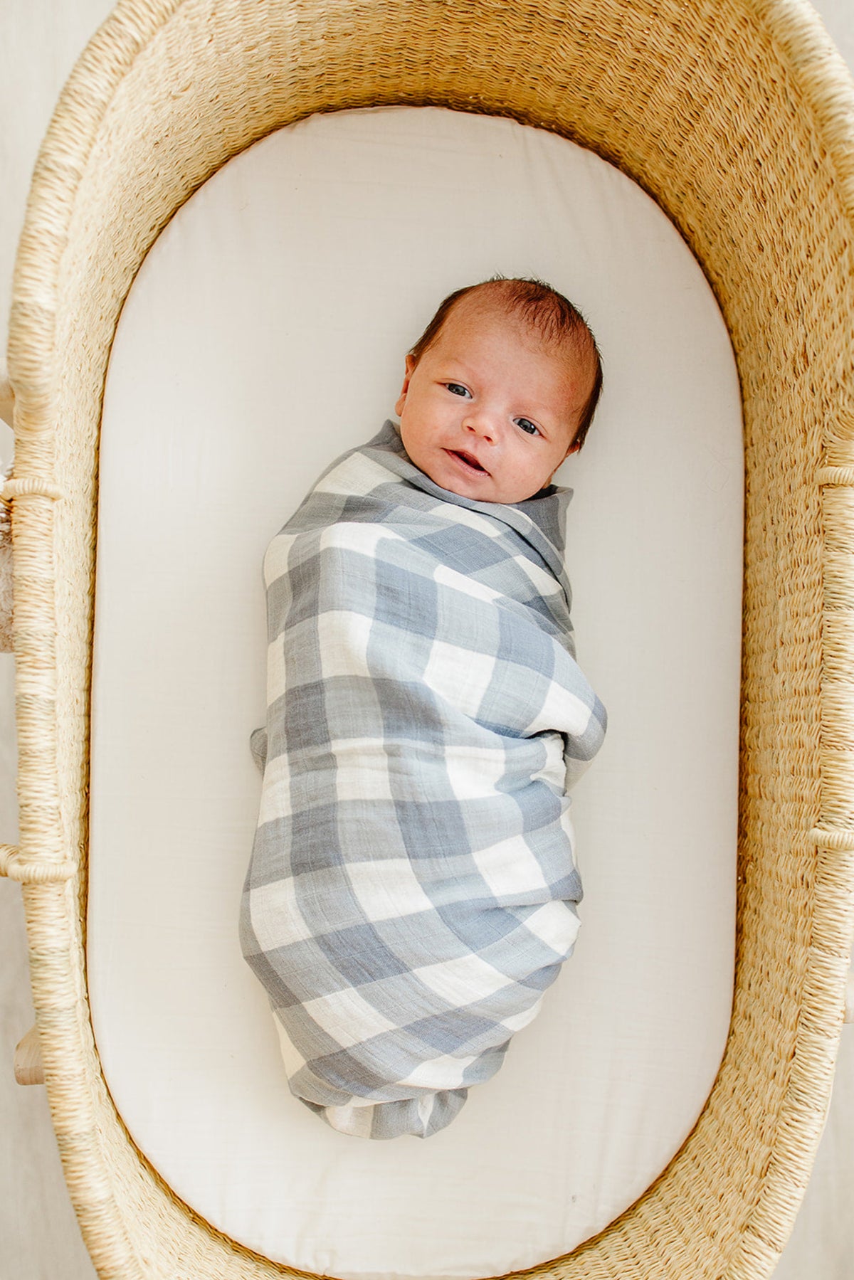 A baby is wrapped in a dusty blue gingham muslin swaddle blanket, lying in a wicker bassinet.
