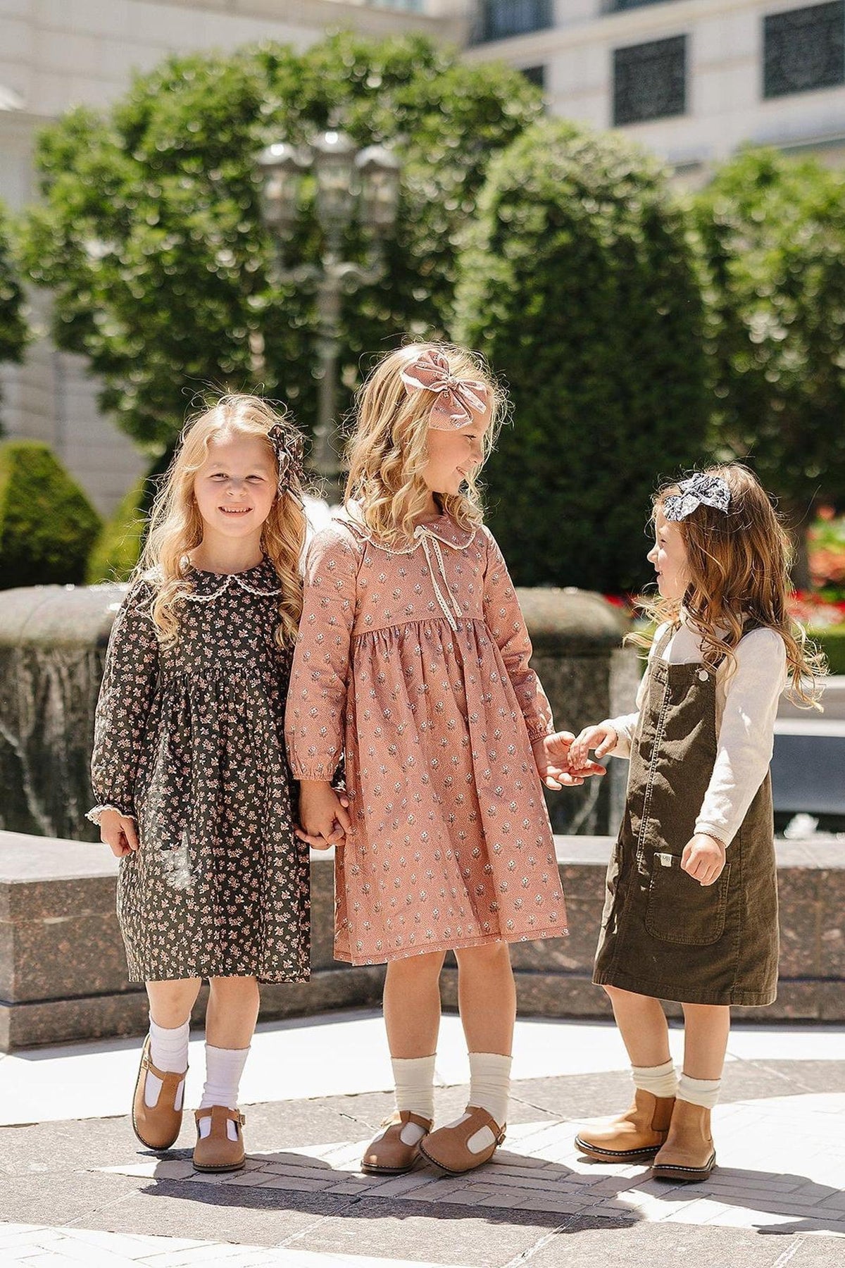 Three young girls wearing floral and solid dresses, standing outdoors near a fountain, smiling and holding hands.