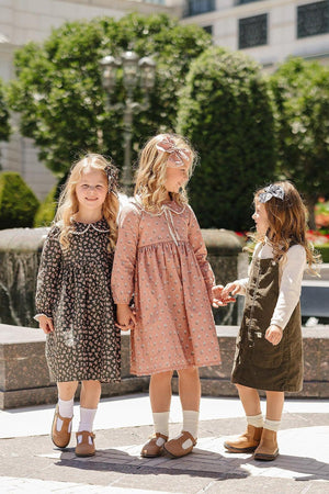 Three young girls wearing floral and solid dresses, standing outdoors near a fountain, smiling and holding hands.