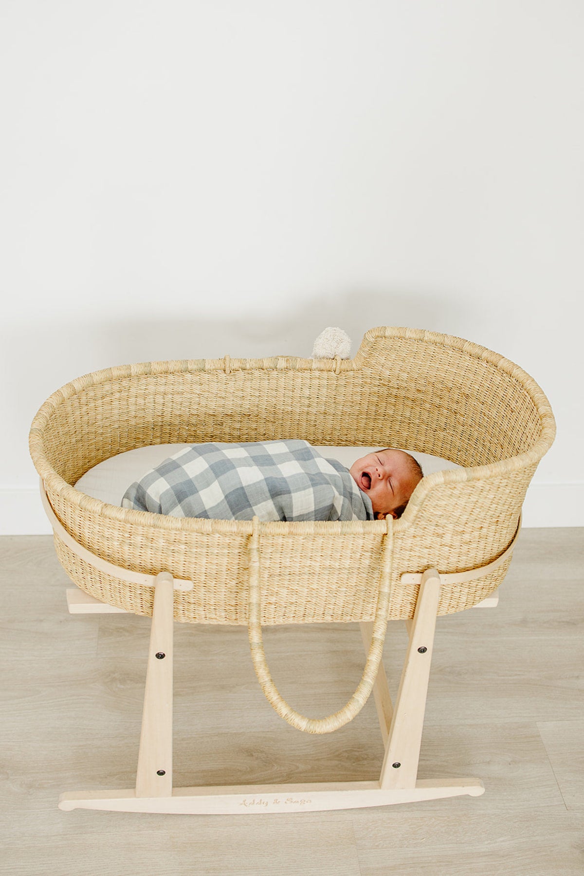 A baby is swaddled in a dusty blue gingham blanket, resting in a woven bassinet on a wooden stand.