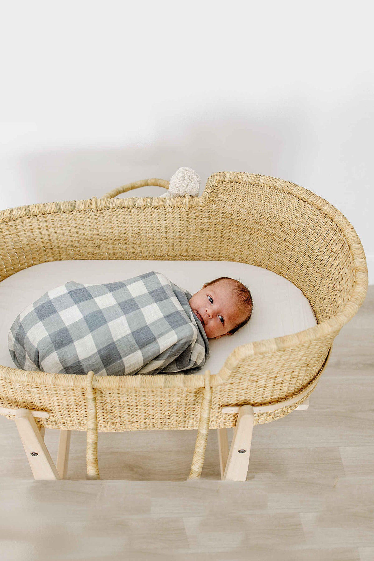 A baby is swaddled in a dusty blue gingham blanket, lying in a woven bassinet on a light floor.
