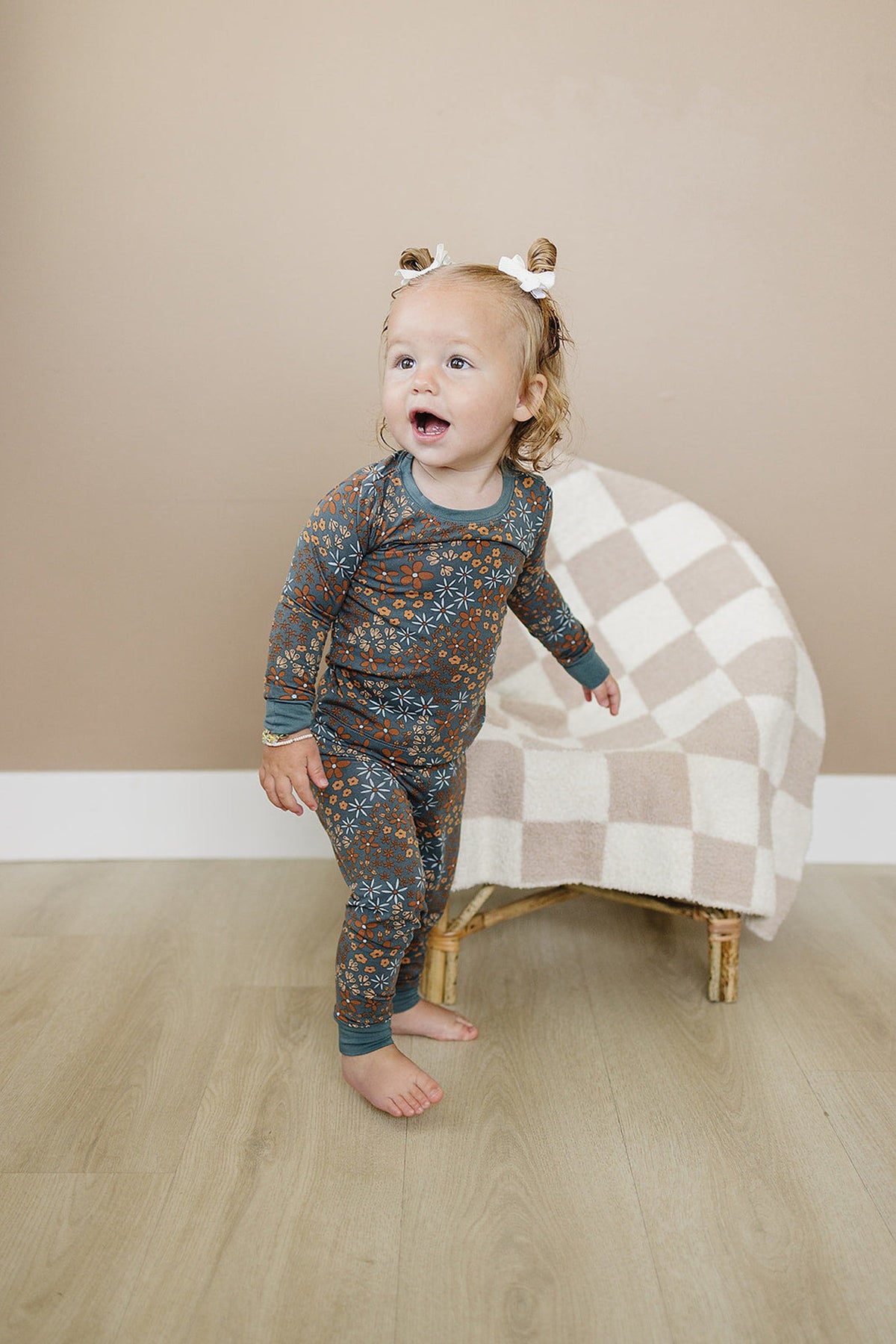 A toddler in a navy floral pajama set stands on a wooden floor beside a checkered blanket-covered chair.