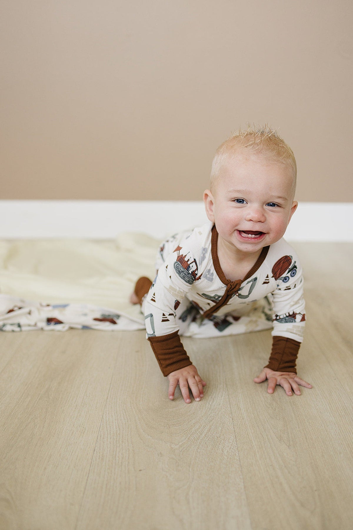 A baby crawls on wooden flooring, wearing a construction truck patterned pajama with brown cuffs, smiling cheerfully.
