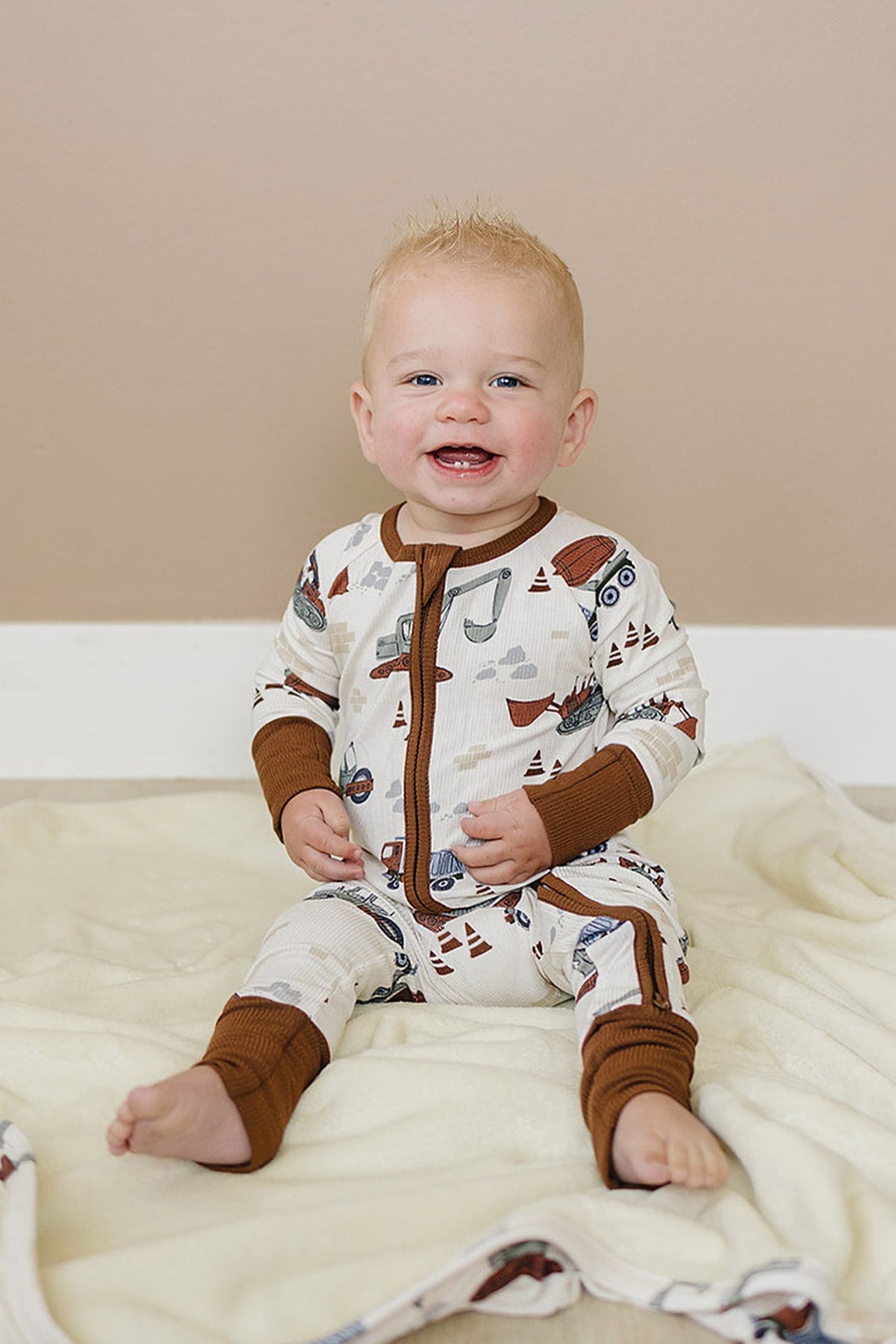 Smiling baby wearing a ribbed pajama with construction truck pattern, featuring brown trim, sitting on a cream blanket.