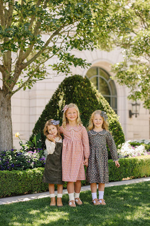 Three young girls in floral dresses stand smiling in a garden with lush greenery and flowers.