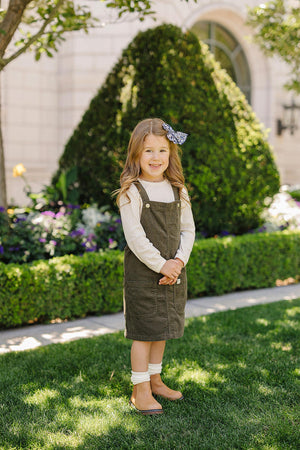A young girl in a green corduroy dress stands smiling in a garden, wearing a bow in her hair.