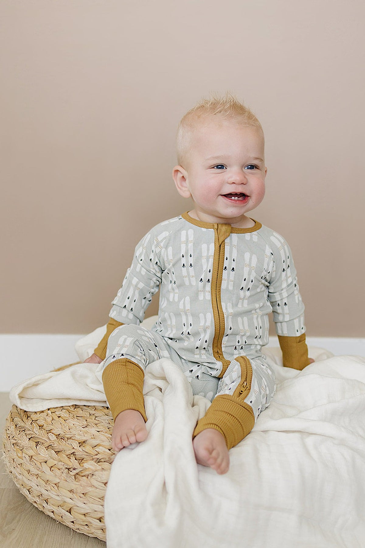Smiling baby wearing a patterned, ribbed pajama with mustard cuffs, sitting on a woven cushion with a white blanket.