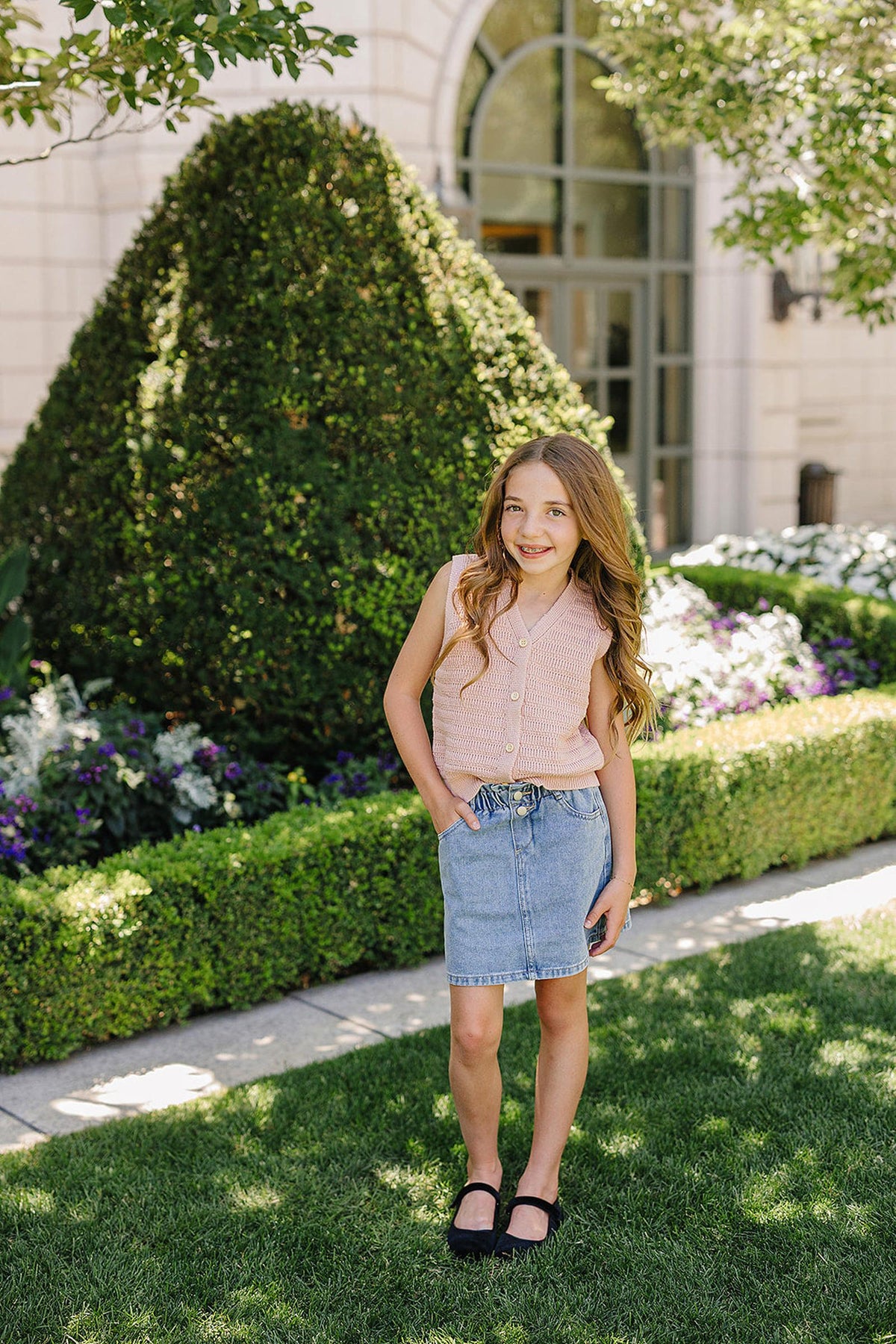 Young girl wearing a pink knit button-down vest and denim skirt, standing in a lush garden setting.