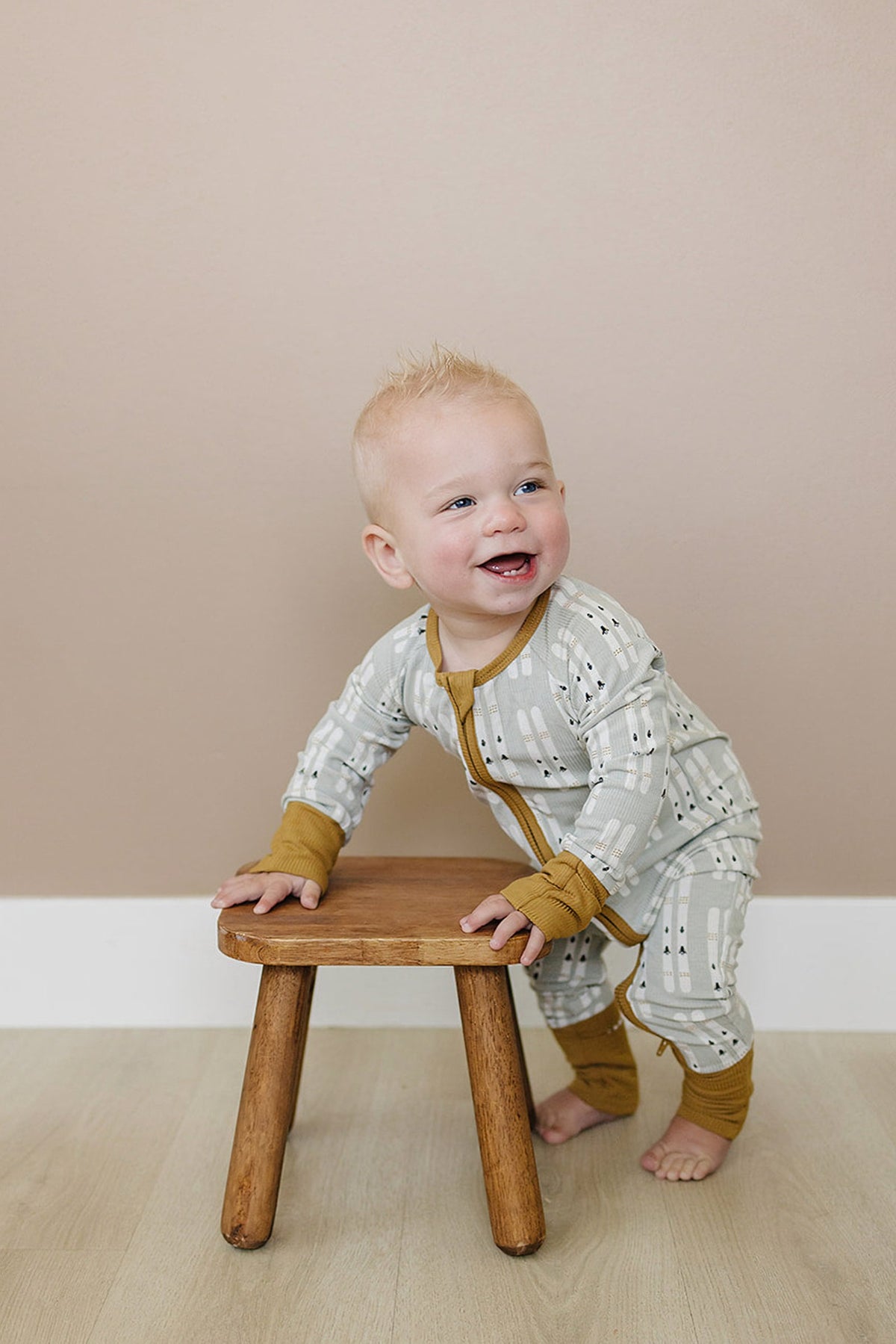 A smiling baby in a ribbed, ski-patterned pajama stands by a wooden stool on a light floor.
