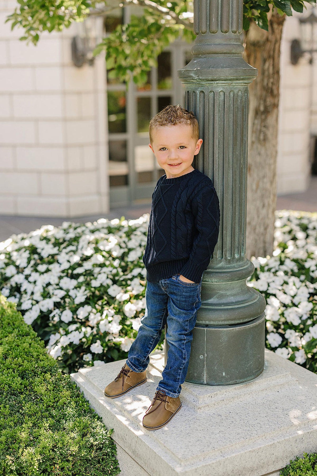 A child in a navy cable knit sweater and jeans stands by a lamppost, surrounded by white flowers.