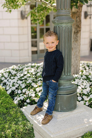 A child in a navy cable knit sweater and jeans stands by a lamppost, surrounded by white flowers.