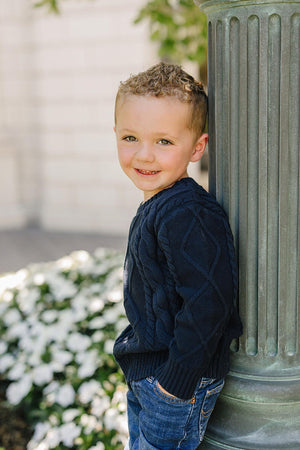 A child in a navy cable knit sweater leans against a pillar, surrounded by greenery and white flowers.