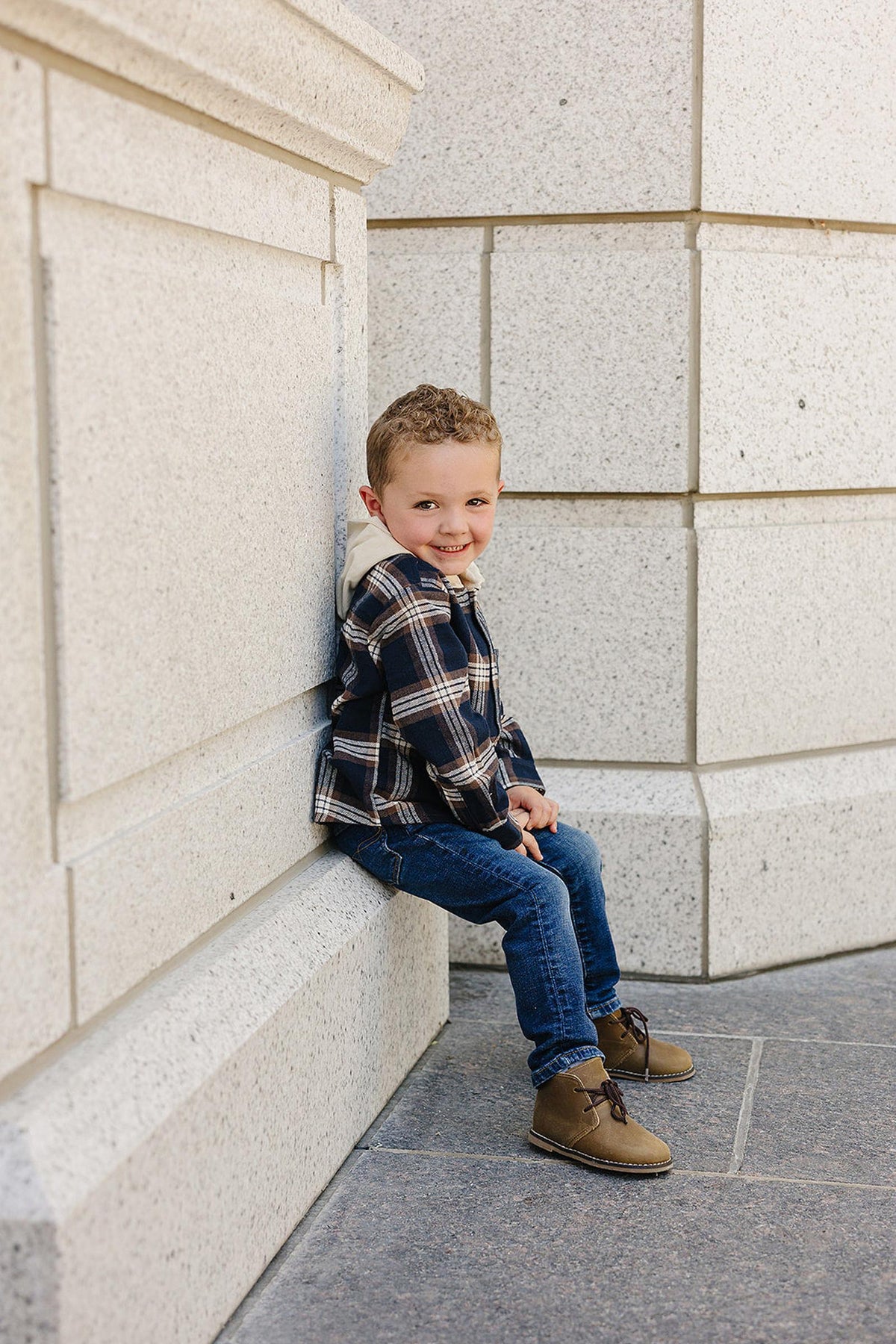 A child wearing a plaid hooded shirt and jeans sits on a stone ledge, smiling outdoors.
