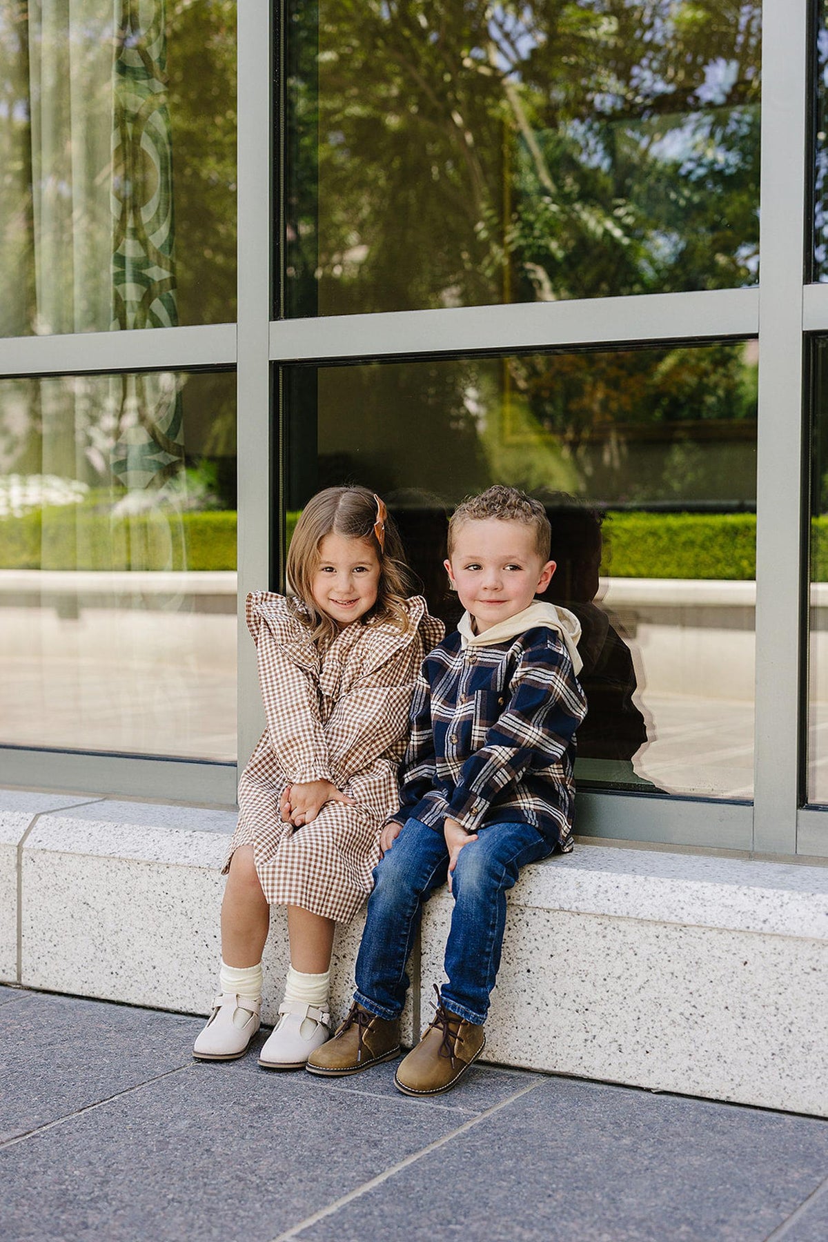 Two children sit outside, wearing plaid outfits; one in a gingham dress, the other in a hooded shirt.
