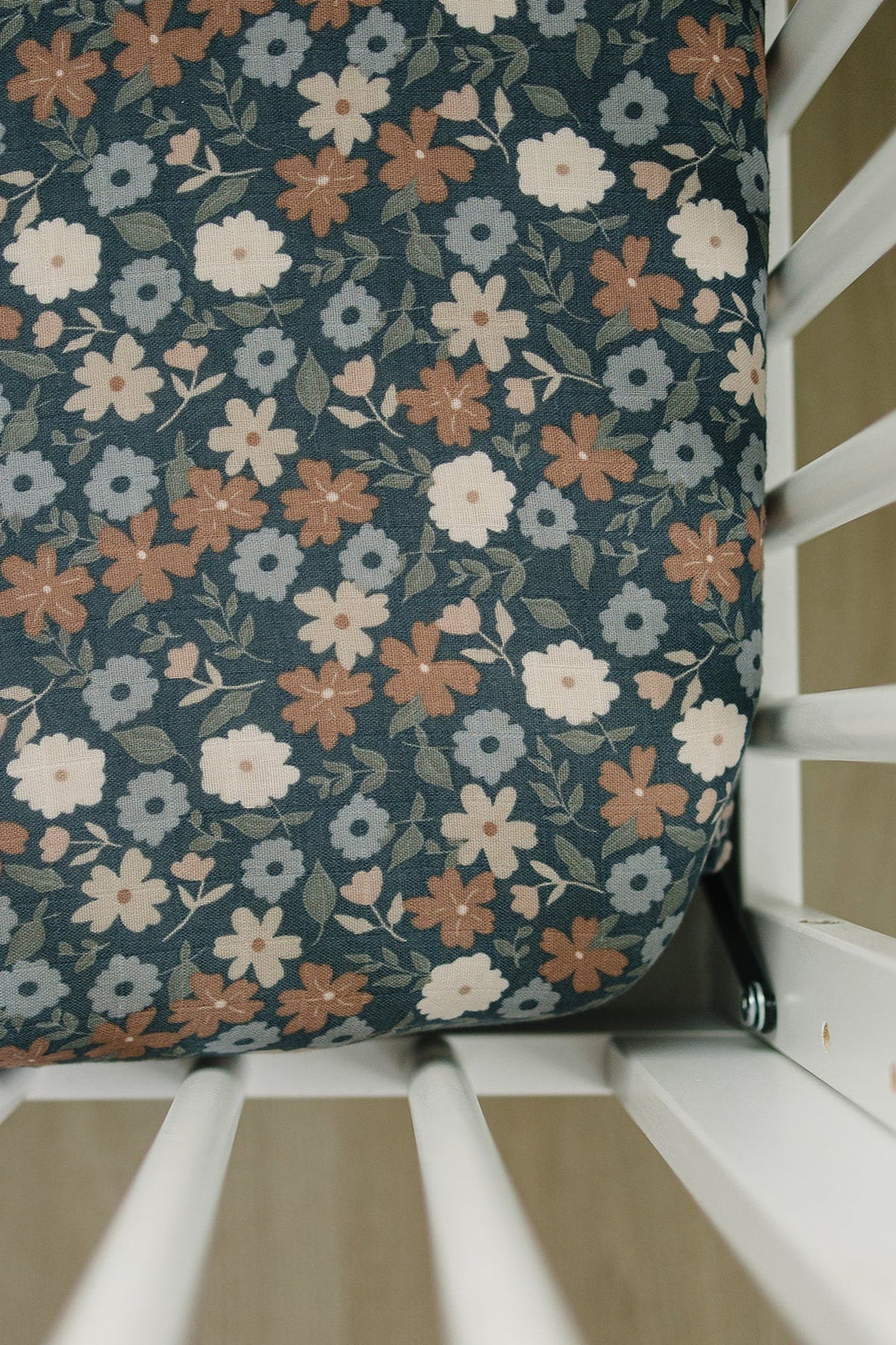 A muslin crib sheet featuring a midnight floral pattern in soft colors of blue, pink, and cream, made from 100% cotton.