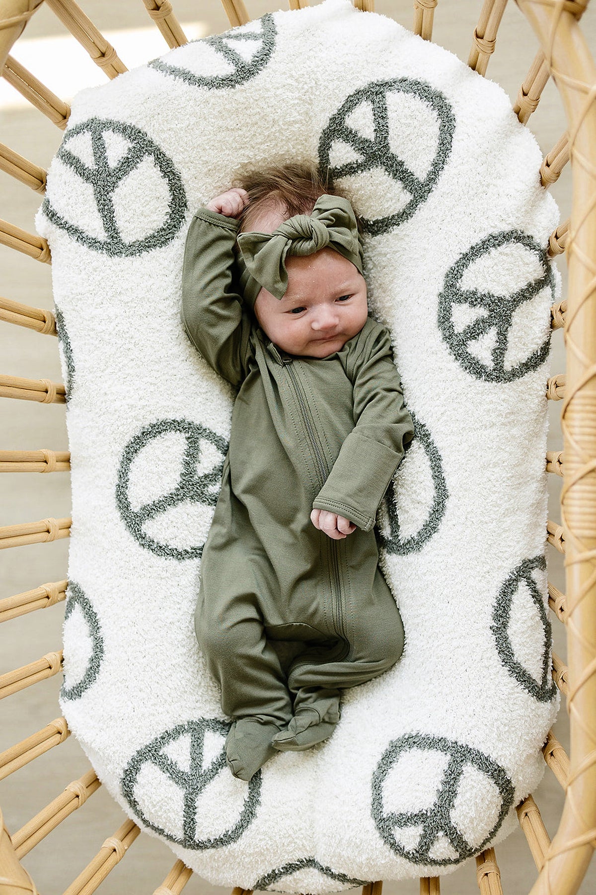 A baby wearing an olive green footed pajama and headband lies on a white cushion with peace signs.