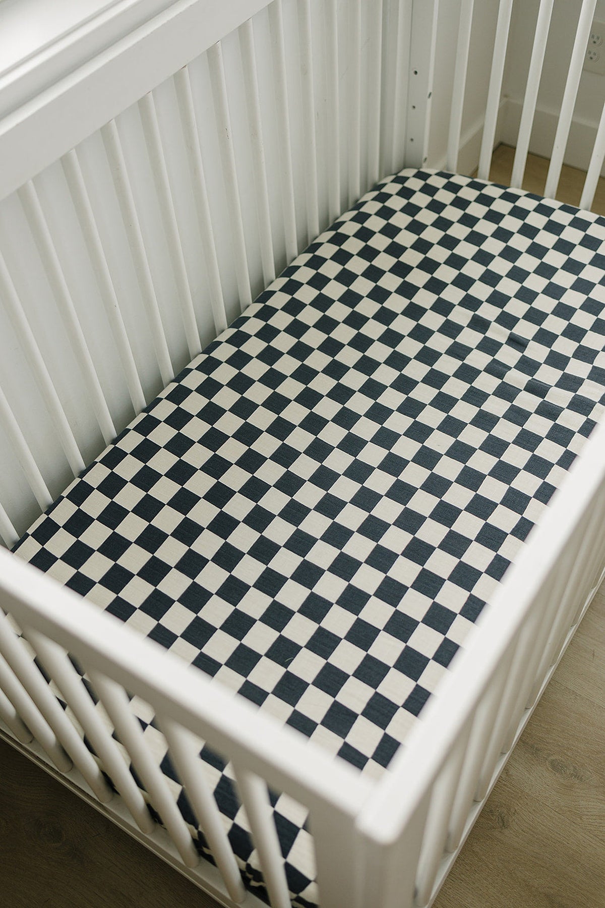 White crib with a charcoal checkered muslin crib sheet, set in a bright nursery with wooden flooring.