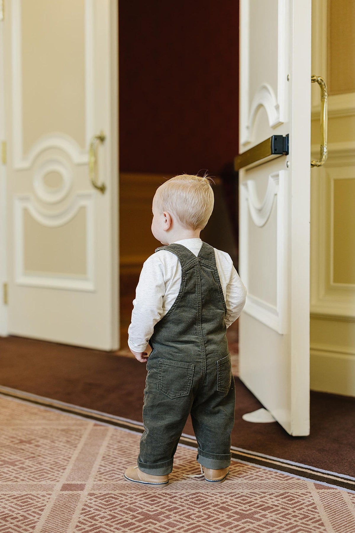 A toddler wearing green corduroy overalls and a white shirt stands near an open door, facing away.