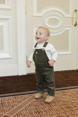 A toddler wearing green corduroy overalls and a white shirt stands on a patterned carpet indoors.